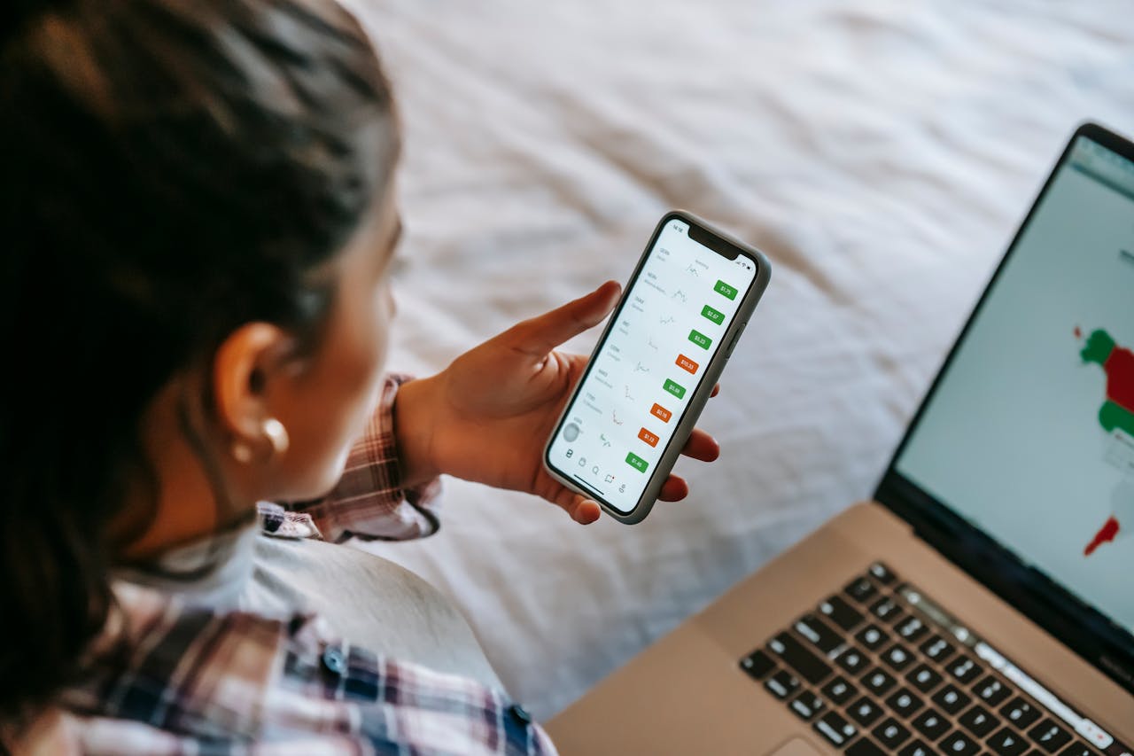 Female analyzing stock market data on her smartphone and laptop indoors. Ideal for finance, technology, and investment themes.